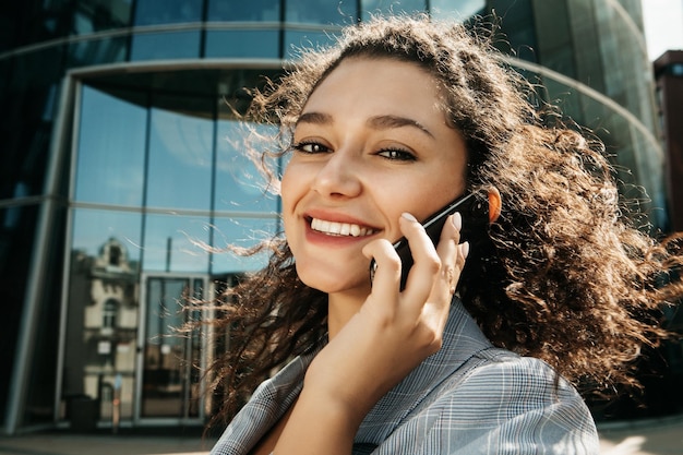 Uma jovem afro-americana vestida em estilo de negócios no contexto de um centro de negócios está segurando um telefone celular e falando