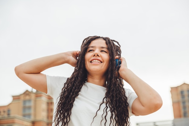 Foto uma jovem alegre e feliz com dreadlocks vestido com uma camiseta branca dançando ouvindo música com fones de ouvido descansando relaxando em um parque da cidade caminhando por um beco conceito de estilo de vida urbano