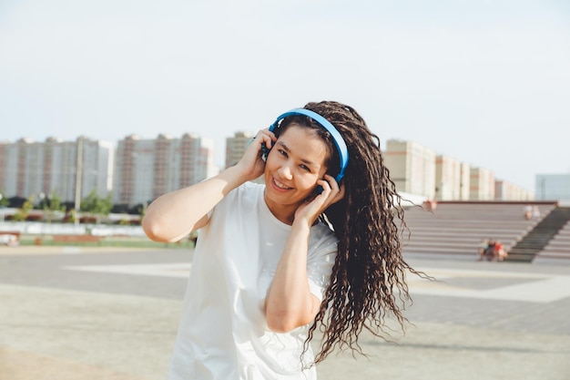 Uma jovem alegre e feliz com dreadlocks vestido com uma camiseta branca dançando ouvindo música com fones de ouvido descansando relaxando em um parque da cidade caminhando por um beco conceito de estilo de vida urbano