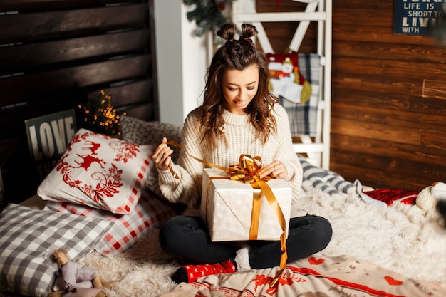 Uma jovem alegre e feliz em um suéter vintage de tricô abre um presente em uma cama na véspera de Natal