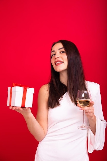 Uma jovem alegre em um vestido branco posa em um fundo vermelho com um copo de vinho e uma caixa com um presente para o ano novo A atmosfera do feriado Fotografia vertical