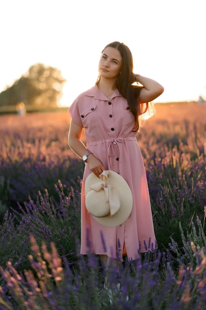 Uma jovem alegre em um vestido rosa e um chapéu de palha nas mãos fica entre arbustos de lavanda