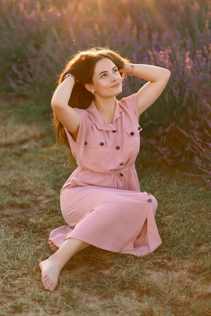 Uma jovem alegre em um vestido rosa e um chapéu de palha nas mãos fica entre arbustos de lavanda