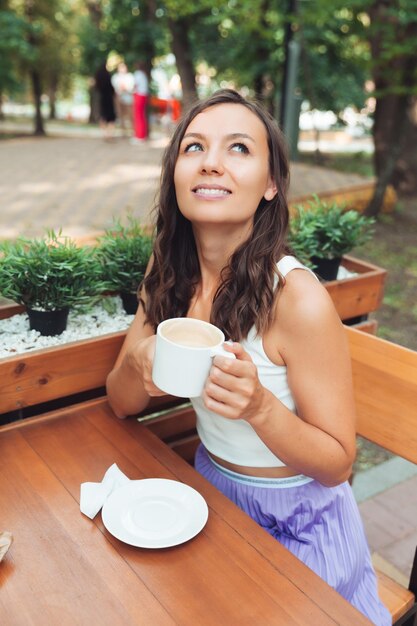 Uma jovem alegre está sentada em um café de verão ao ar livre e bebendo uma bebida