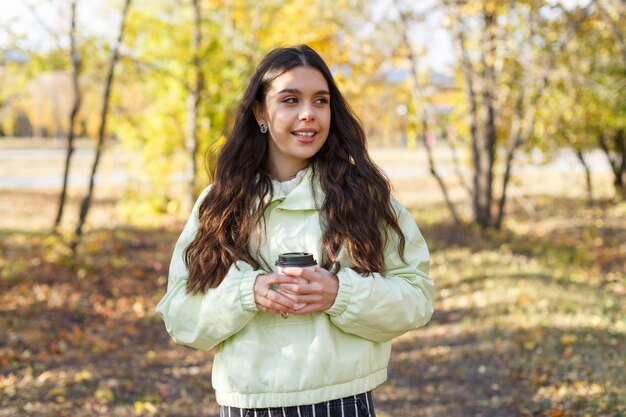 Uma jovem alegre tomando uma xícara de café em um parque no outono