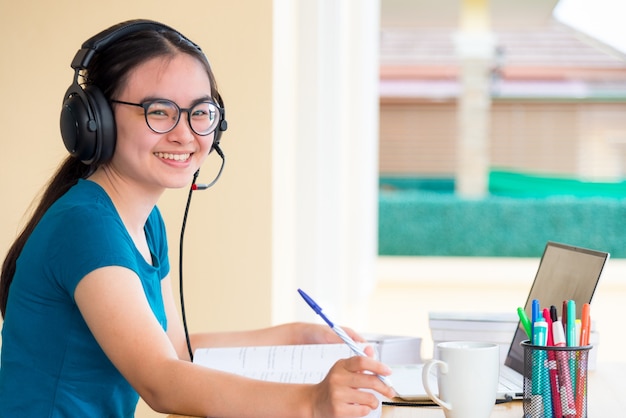 Uma jovem aluna adolescente asiática com óculos de fone de ouvido está olhando enquanto usa um laptop, um estudo on-line de uma escola de educação à distância, aprendendo de uma classe de uma universidade em casa para segundo plano