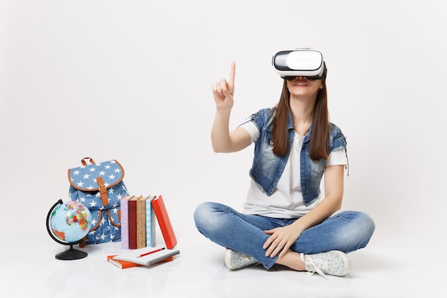 Uma jovem aluna com óculos de realidade virtual toca algo como apertar o botão, apontando para uma tela virtual flutuante perto do globo, mochila, livro escolar isolado