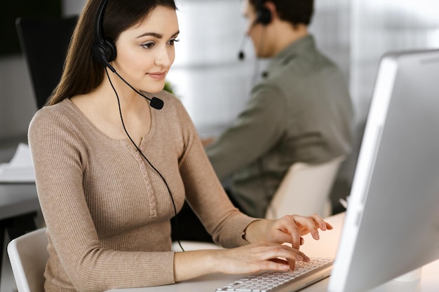 Foto uma jovem amiga com fones de ouvido está a falar com um cliente de uma empresa, sentada à secretária num escritório moderno com um colega. operadores de call center a trabalhar.