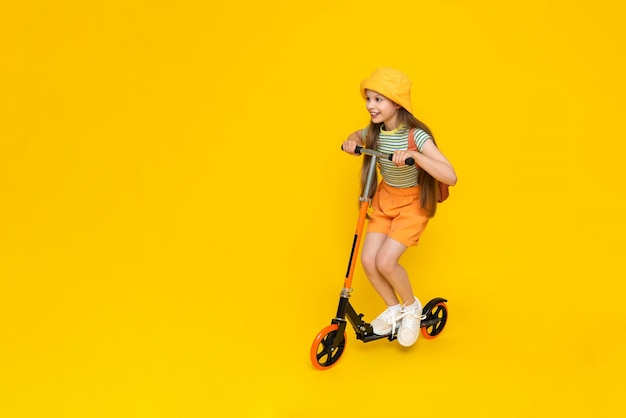 Uma jovem anda de scooter Uma criança feliz em shorts e um chapéu com uma mochila está aproveitando as férias de verão Recreação infantil ativa Fundo isolado amarelo Espaço para cópia