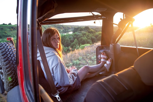 Uma jovem aprecia o pôr do sol enquanto está sentado no porta-malas de um suv