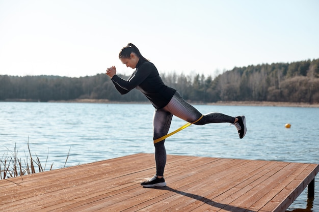 Uma jovem apto realiza exercícios com faixas elásticas de aptidão em um píer na margem do lago. O conceito de um estilo de vida saudável. Vista lateral