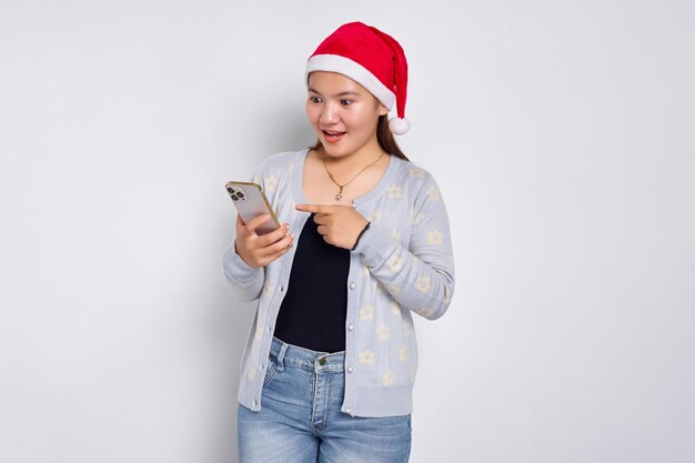 Uma jovem asiática alegre com um chapéu de Natal apontando dedos para o celular isolado sobre fundo branco