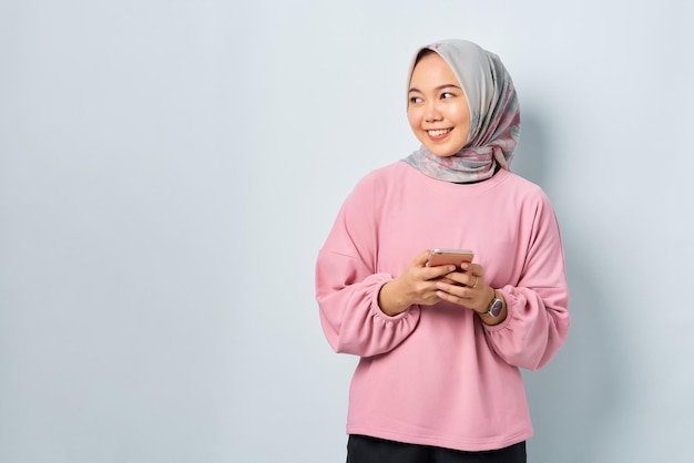 Uma jovem asiática alegre de camisa rosa usando telefone celular e desviando o olhar para o espaço em branco isolado sobre fundo branco