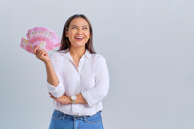 Uma jovem asiática alegre segurando notas de dinheiro e olhando para o espaço de cópia isolado sobre fundo branco
