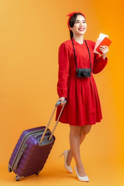 Uma jovem asiática alegre segurando passaporte de mala e passagem de avião enquanto estava isolada sobre fundo laranja
