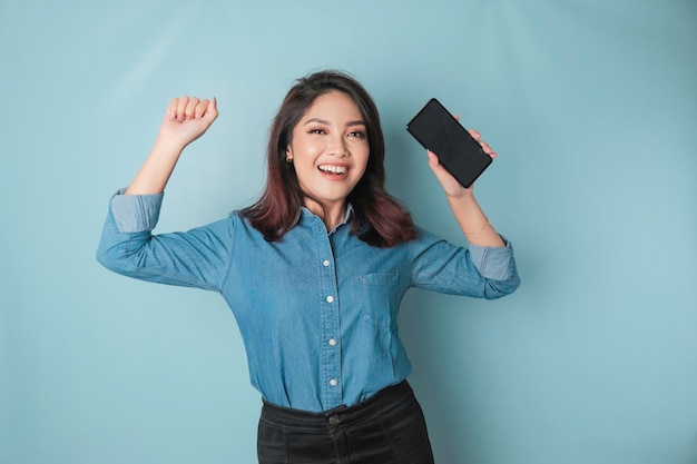 Uma jovem asiática com uma expressão feliz e bem-sucedida vestindo uma camisa azul segurando smartphone isolado por fundo azul