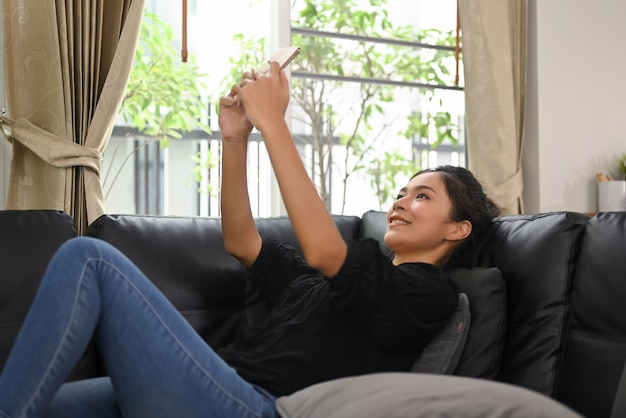 Uma jovem asiática está usando um telefone celular enquanto está deitada no sofá de couro preto