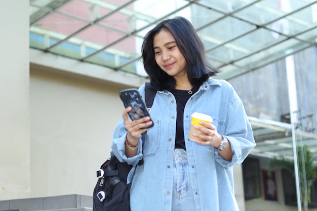 uma jovem asiática excitada está rindo enquanto usa um aplicativo em um smartphone e bebe café nas escadas exteriores de um shopping