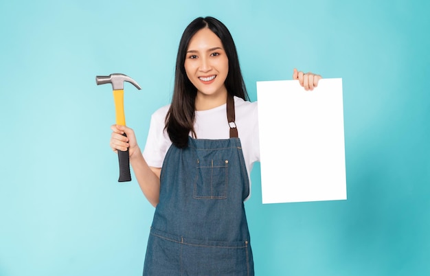 Foto uma jovem asiática feliz usa um avental e segura um martelo de carpinteiro mostrando um papel em branco sobre fundo azul