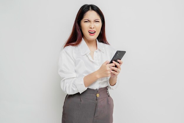 Uma jovem asiática insatisfeita parece descontente vestindo camisa branca expressões faciais irritadas segurando seu telefone