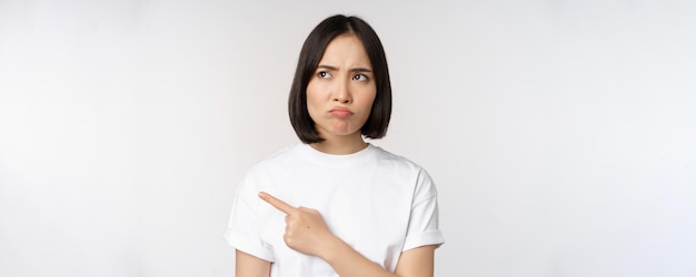 Uma jovem asiática irritada e chateada apontando para a esquerda com arrependimento ou ciúme se sente injusto em pé sobre fundo branco