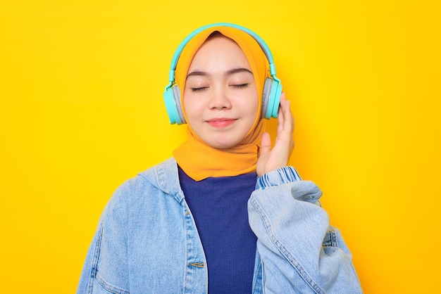 Uma jovem asiática relaxada em jaqueta jeans usando fones de ouvido para ouvir música isolada sobre fundo amarelo