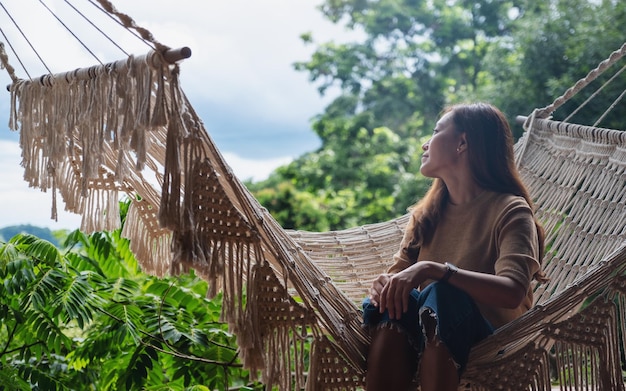 Uma jovem asiática sentada e relaxando na rede enquanto olha para uma bela vista da natureza