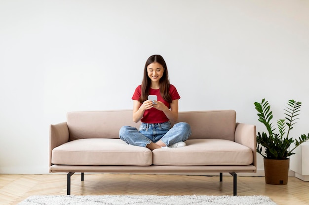 Uma jovem asiática sorridente com um smartphone a relaxar no sofá em casa.