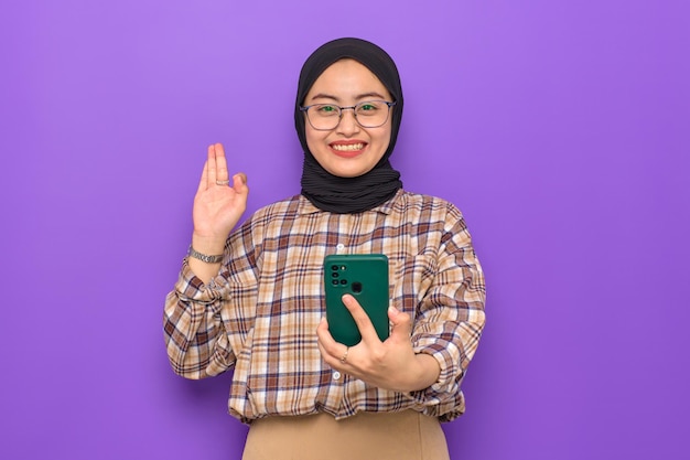 Uma jovem asiática sorridente em camisa xadrez segurando o celular e mostrando um gesto bem isolado em fundo roxo