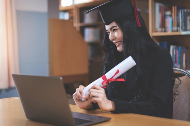 Uma jovem asiática universitária expressando alegria e entusiasmo ao comemorar a conquista da formatura em frente a um laptop, fazendo uma videochamada remota para os pais em casa