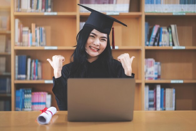 Uma jovem asiática universitária expressando alegria e entusiasmo ao comemorar a conquista da formatura em frente a um laptop, fazendo uma videochamada remota para os pais em casa