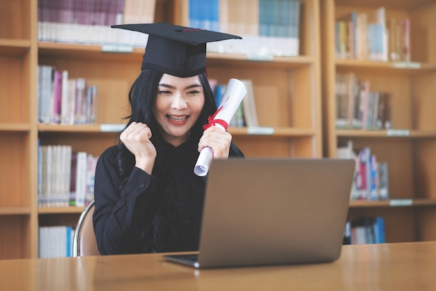 Uma jovem asiática universitária expressando alegria e entusiasmo ao comemorar a conquista da formatura em frente a um laptop, fazendo uma videochamada remota para os pais em casa