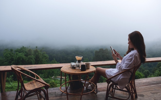 Uma jovem asiática usando telefone celular enquanto está sentado na varanda com uma bela vista da natureza em dia de neblina