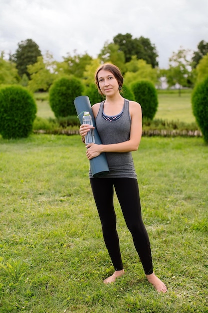 Uma jovem atlética em um agasalho cinza para fitness vai fazer ioga em um parque verde.