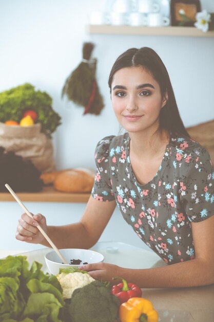 Uma jovem atraente de cabelos escuros prova uma nova receita para uma deliciosa mistura de salada enquanto está sentada à mesa na cozinha