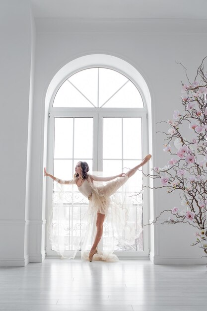 uma jovem bailarina esbelta está dançando em uma sala iluminada perto da janela em um vestido