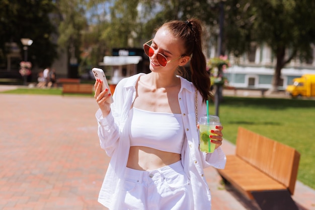 Uma jovem bebe um coquetel refrescante de verão na cidade e olha para o telefone