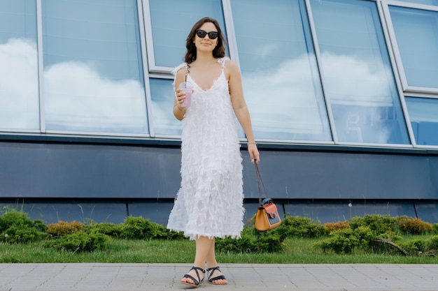 Uma jovem bebe um smoothie do lado de fora Roupa elegante, vestido branco, óculos de sol e bolsa Caminhada de verão