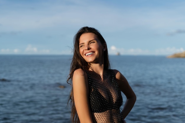 Uma jovem bela mulher bronzeada contra o pano de fundo do mar ao pôr do sol em um maiô preto e um vestido de malha ri e sorri com um sorriso branco como a neve