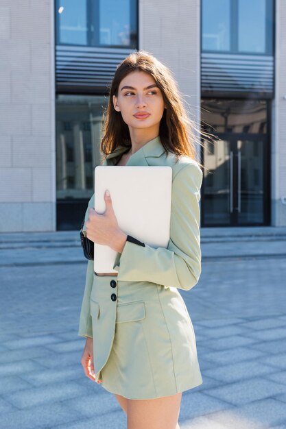 Uma jovem bem-sucedida em um terno estiloso está segurando um laptop do lado de fora do retrato comercial