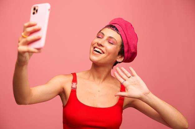 Uma jovem blogueira com cabelo rosa colorido e um corte de cabelo curto tira uma foto de si mesma no telefone e transmite um sorriso em roupas elegantes e um chapéu em um estilo monocromático de fundo rosa