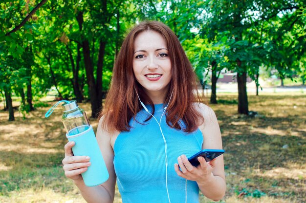 Uma jovem bonita com cabelo ruivo olha para a câmera, coloca os fones de ouvido, segurando água nas mãos. Uma garota ouvindo um podcast sobre um estilo de vida saudável.