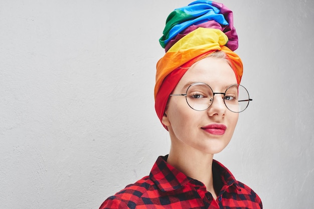 Uma jovem bonita com óculos redondos e lábios carnudos em um turbante de arco-íris e uma camisa vermelha está sorrindo contra uma parede branca. LGBTQ Lésbica, gay, bissexual, transgênero, queer. homem homossexual