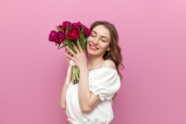 Uma jovem bonita com roupa festiva segura buquê de tulipas cor de rosa e se alegra em fundo rosa