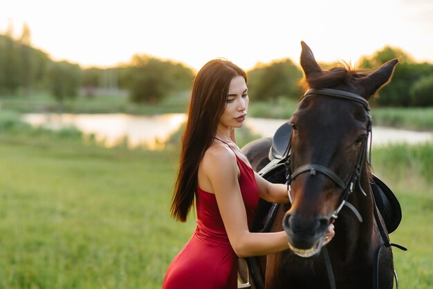 Uma jovem bonita com um vestido vermelho posa em um rancho com um garanhão puro-sangue ao pôr do sol. Amar e cuidar dos animais.