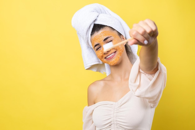 Uma jovem bonita com uma máscara esfoliante no rosto escova os dentes contra um fundo amarelo