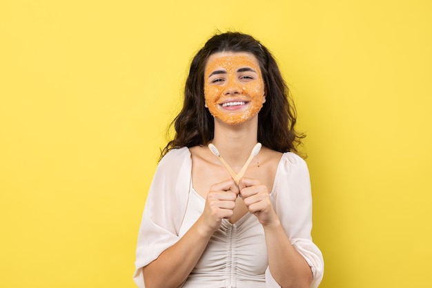 Uma jovem bonita com uma máscara esfoliante no rosto escova os dentes contra um fundo amarelo