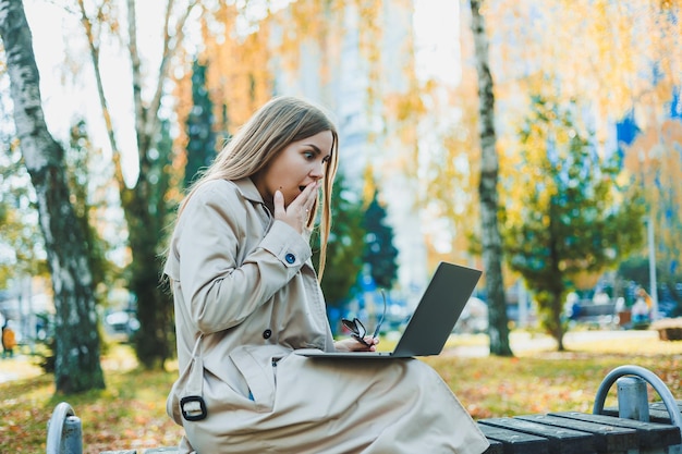 Uma jovem bonita de cabelos louros está sentada em um parque de outono em um banco e trabalhando em um laptop Trabalho remoto