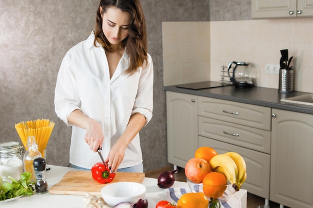 Uma jovem bonita de camisa branca na cozinha corta legumes pimentão vermelho milho cebola azul Vegetarianos comida saudável