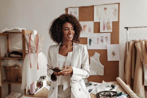 Uma jovem bonita de jaqueta branca de grandes dimensões e calças da moda sorri amplamente segura o telefone Atraente designer de moda posa no escritório